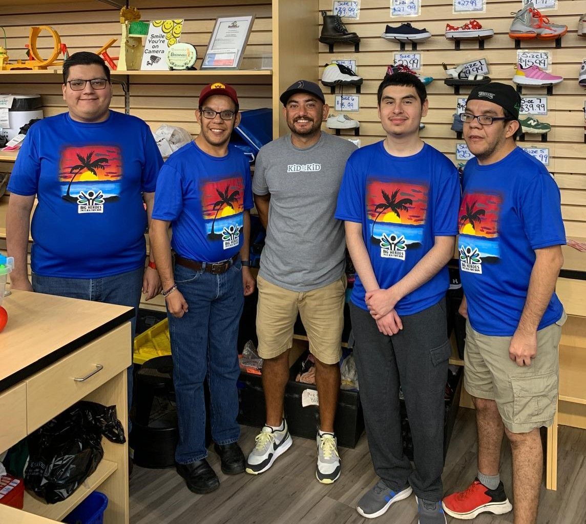 Five people pose in a store. Four wear blue shirts. One wears a grey shirt. Shoes are on the wall behind them.