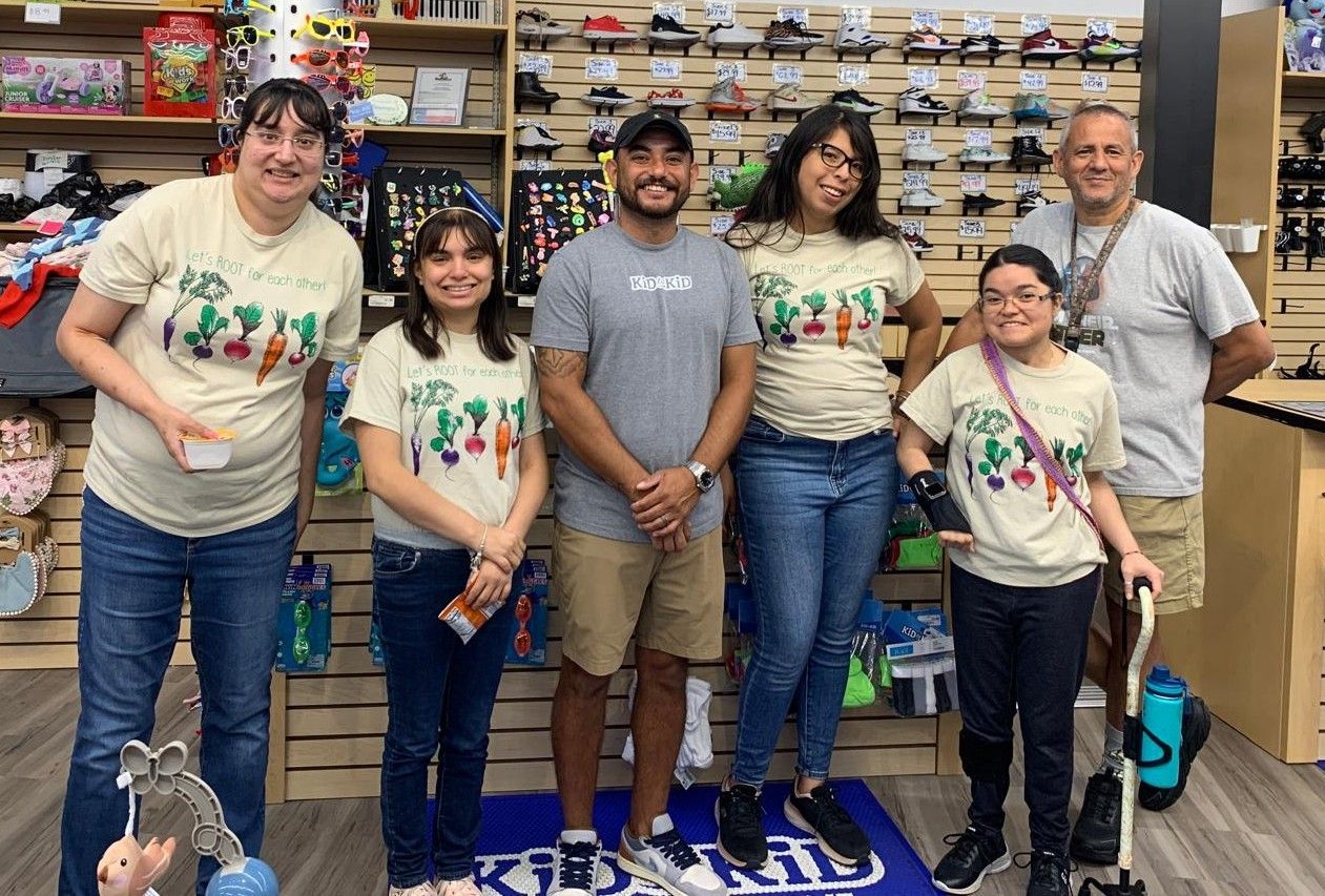 Group of six people smiling, standing inside a skate shop. They are wearing casual clothes, one with a cane.