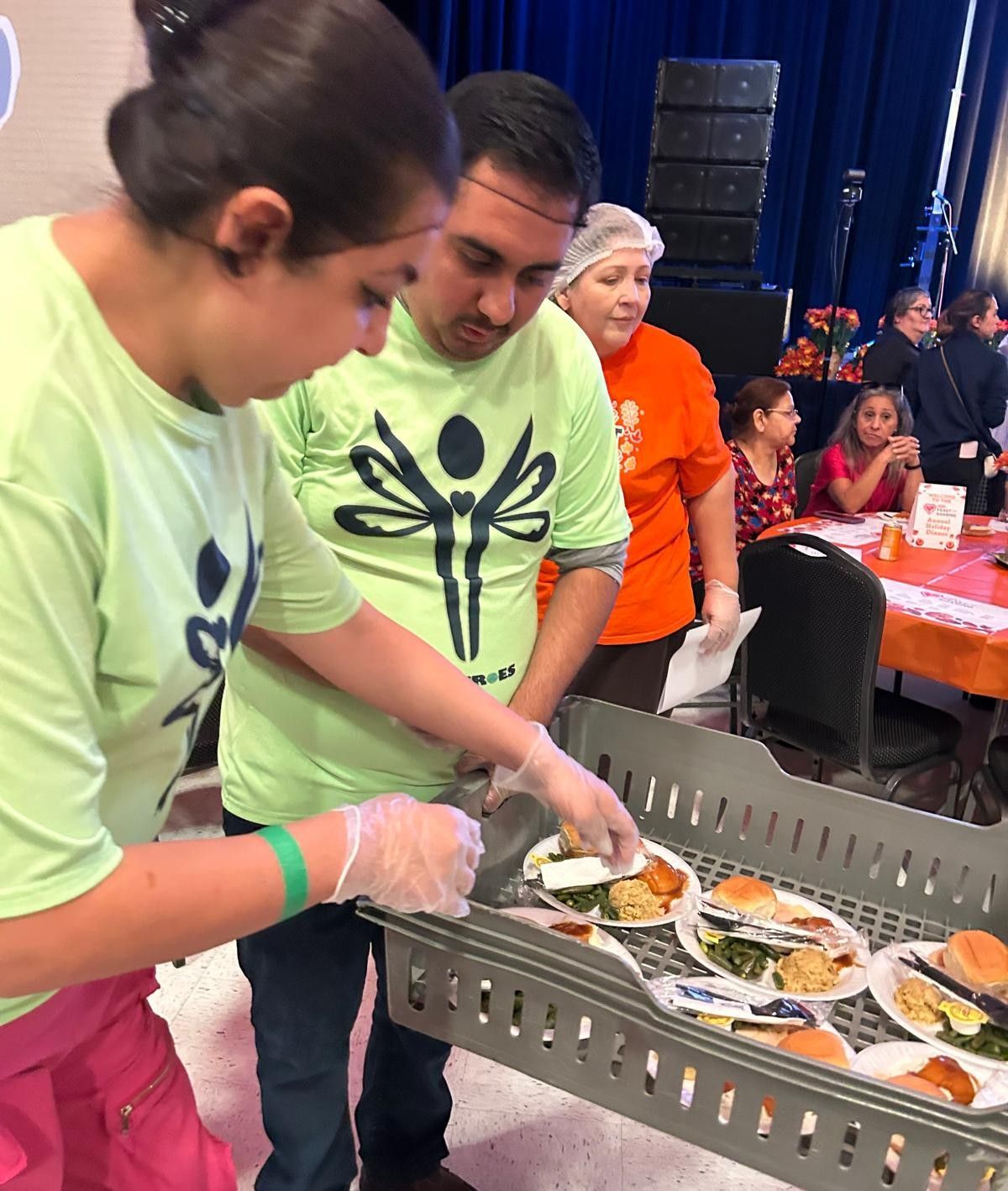 People in green shirts serving food from a tray. Others sit at tables.