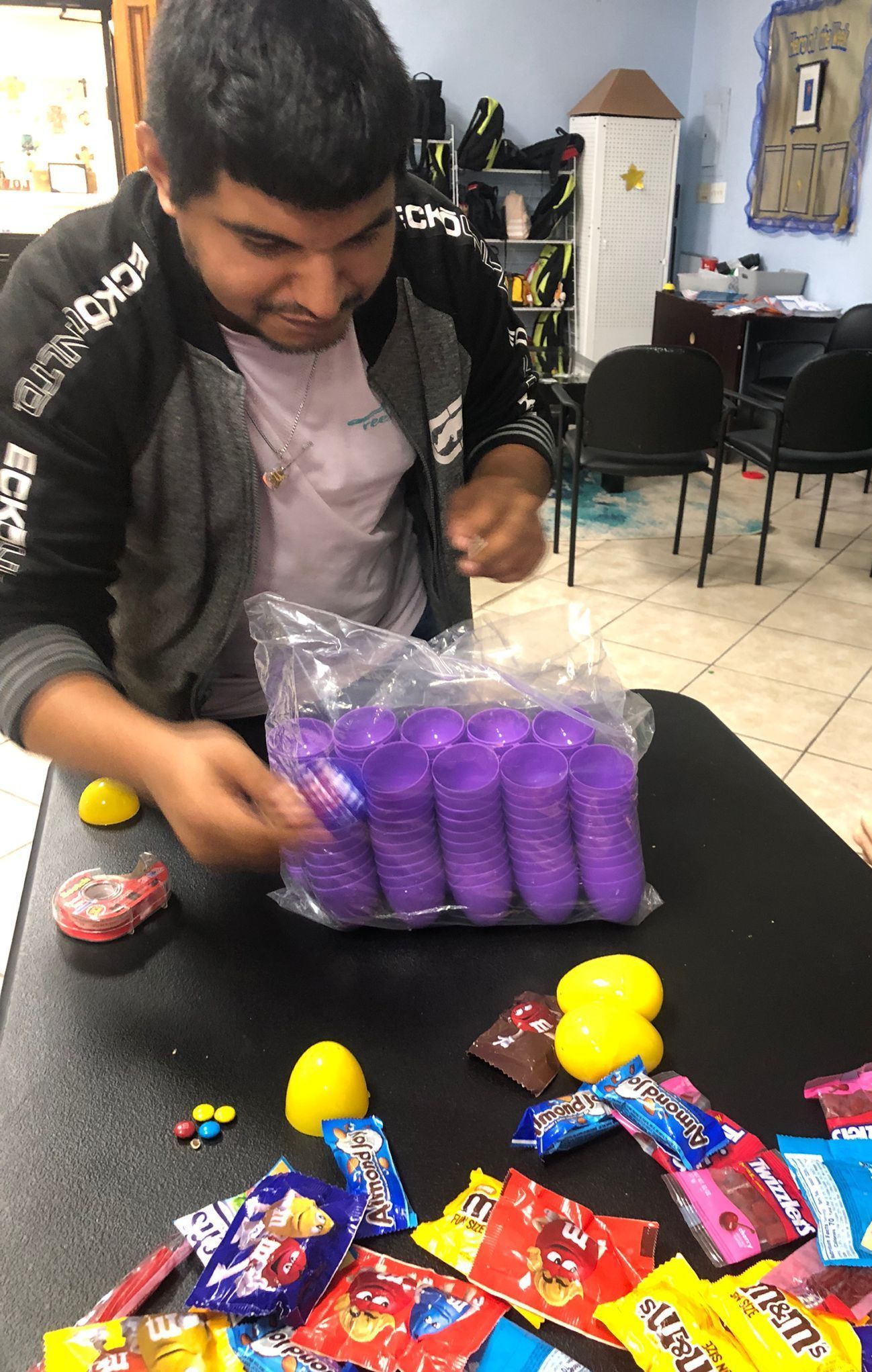 A man is sitting at a table with a bag of purple cups and candy.