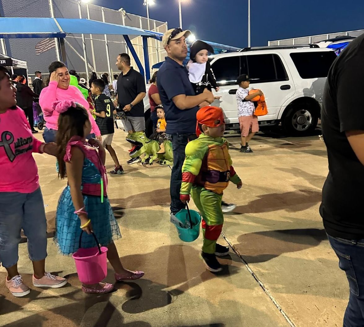 A group of people are standing in a parking lot including a boy in a teenage mutant ninja turtle costume.