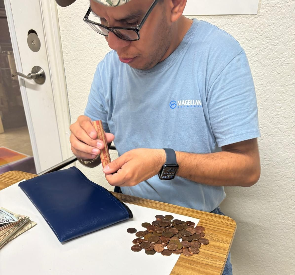 Man counting coins, inserting them into a paper roll at a table. Blue shirt, watch, and glasses.