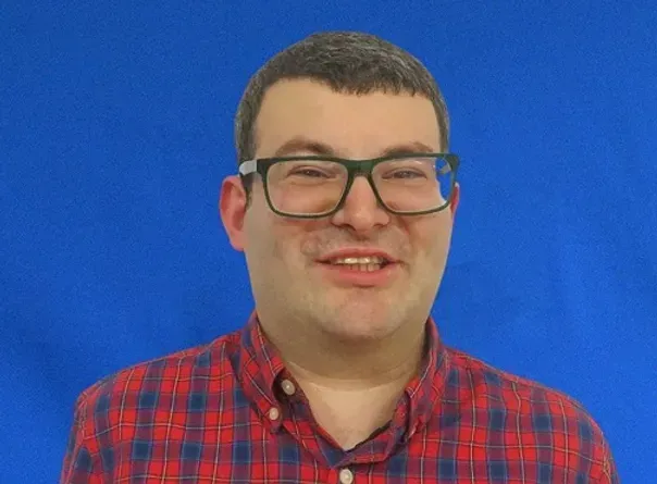 A man wearing glasses and a plaid shirt is smiling in front of a blue background.