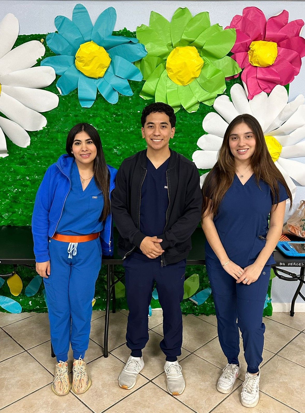 Three people in scrubs are posing for a picture in front of a wall with paper flowers.