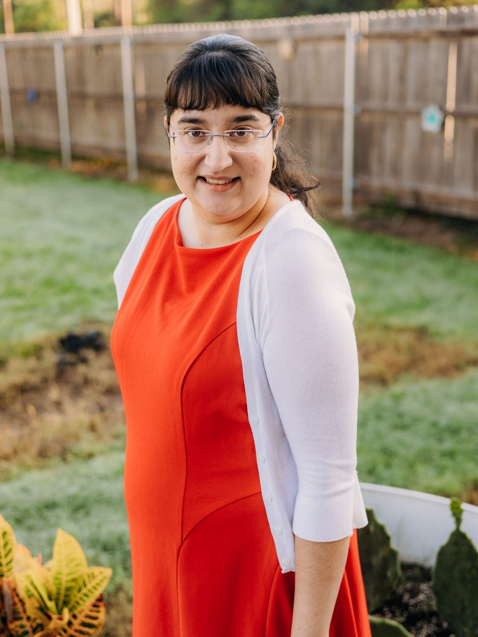 A woman in a red dress and white sweater is standing in front of a fence.