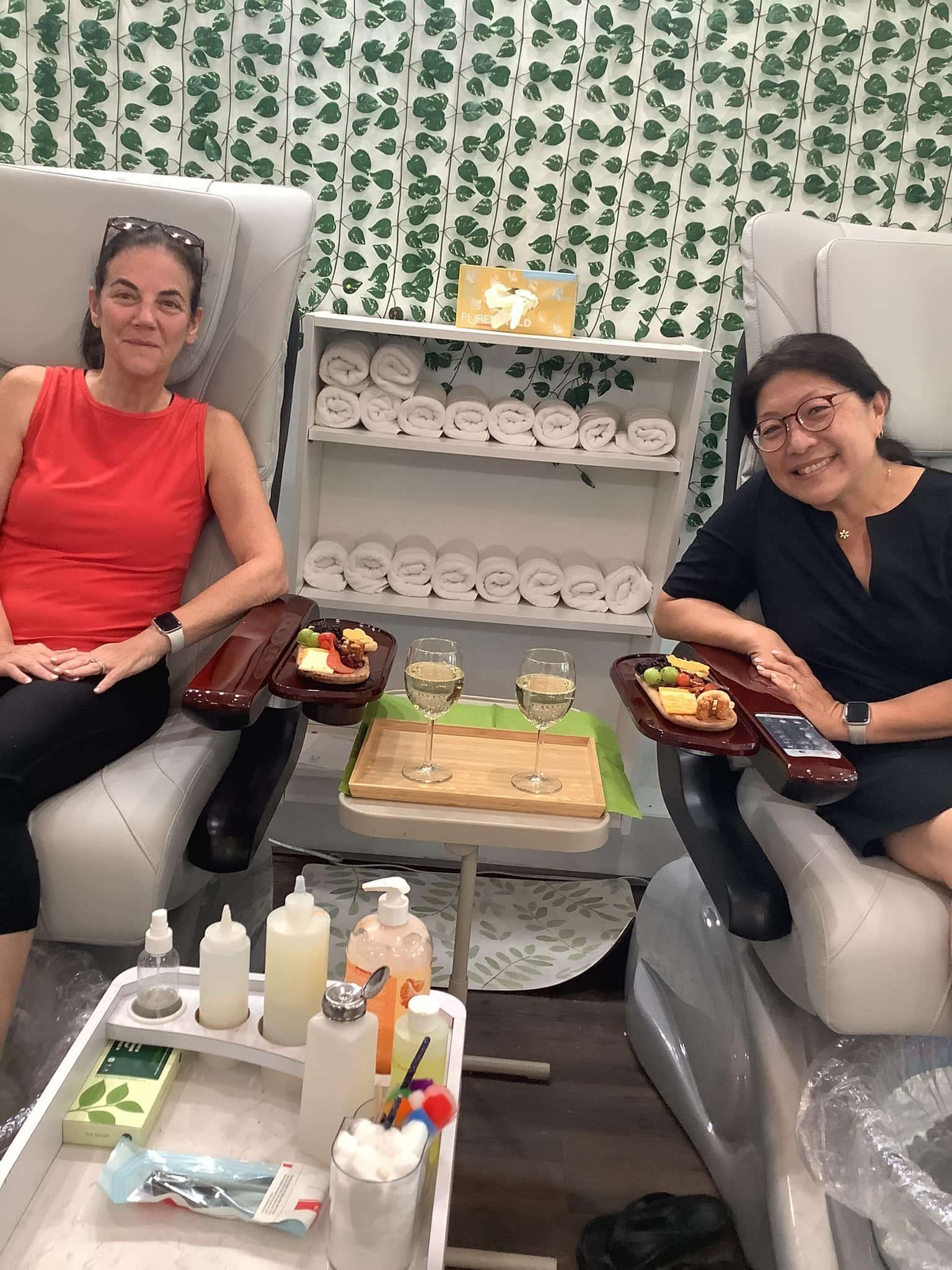 Two women in a nail salon, relaxing in pedicure chairs with drinks and snacks.