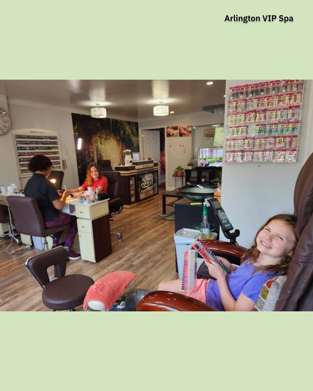 Girl smiles in a nail salon chair as staff works on clients. Wood floors, manicure stations, and nail polish displays.