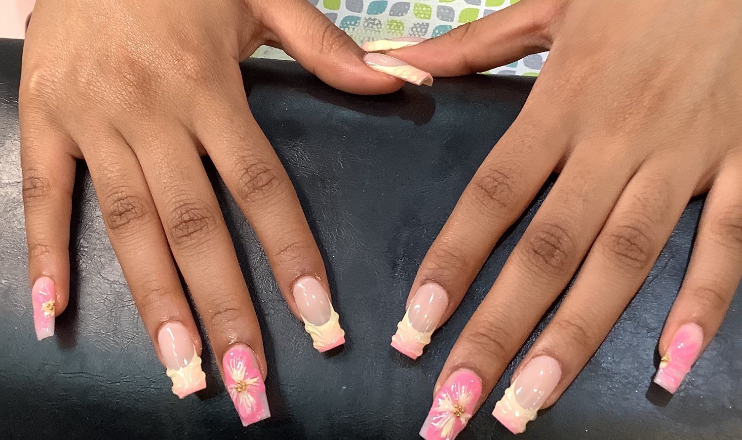 Hands with square, light pink and yellow French tip nails with 3D floral accents.