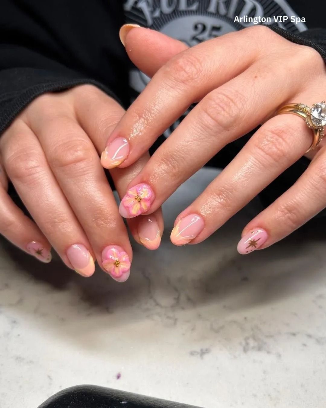 Hands with short, oval-shaped nails. One nail has a pink flower, gold accent. French tip design, gold ring.