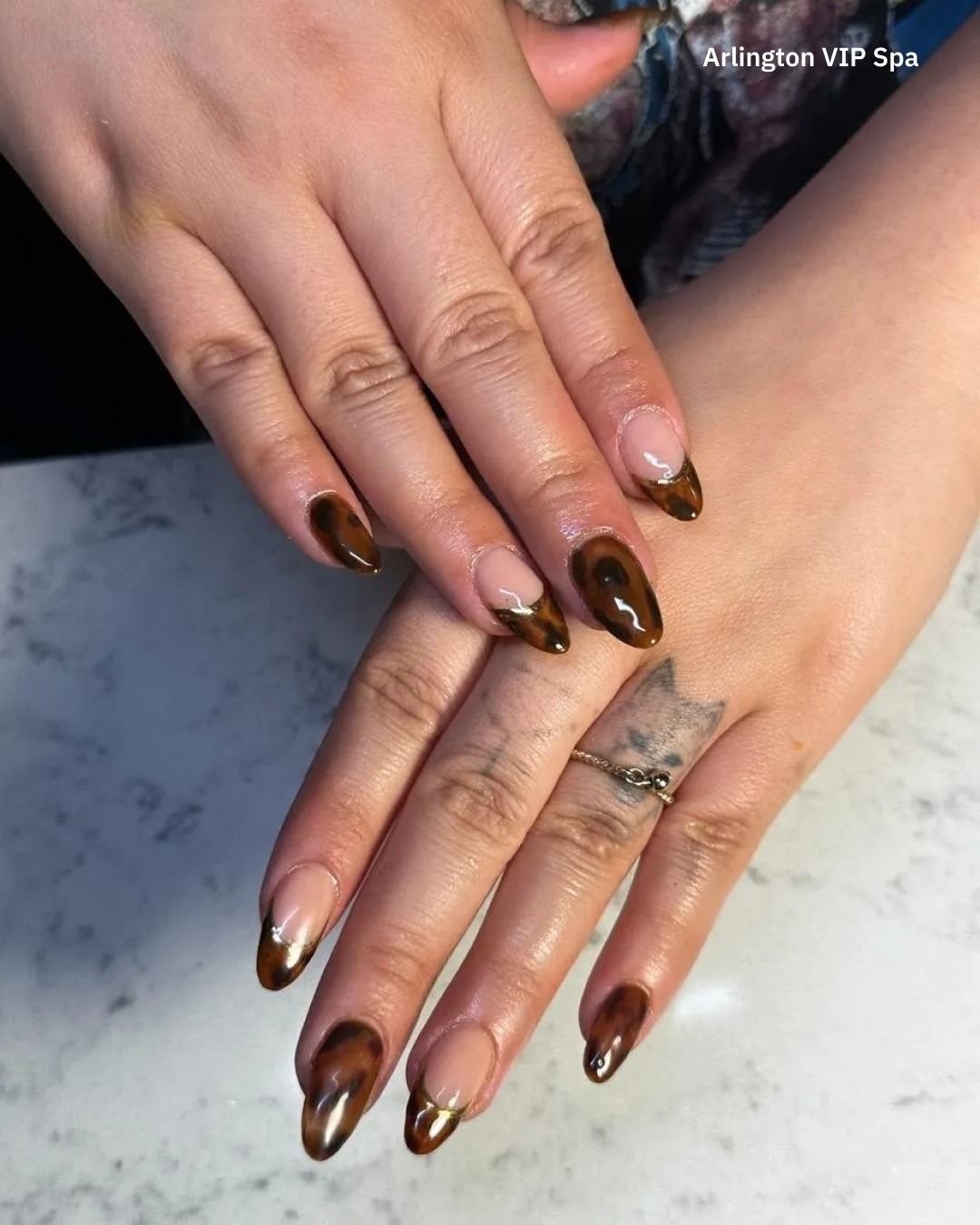 Hands with almond-shaped nails, brown tips, gold trim, and a small cat tattoo.