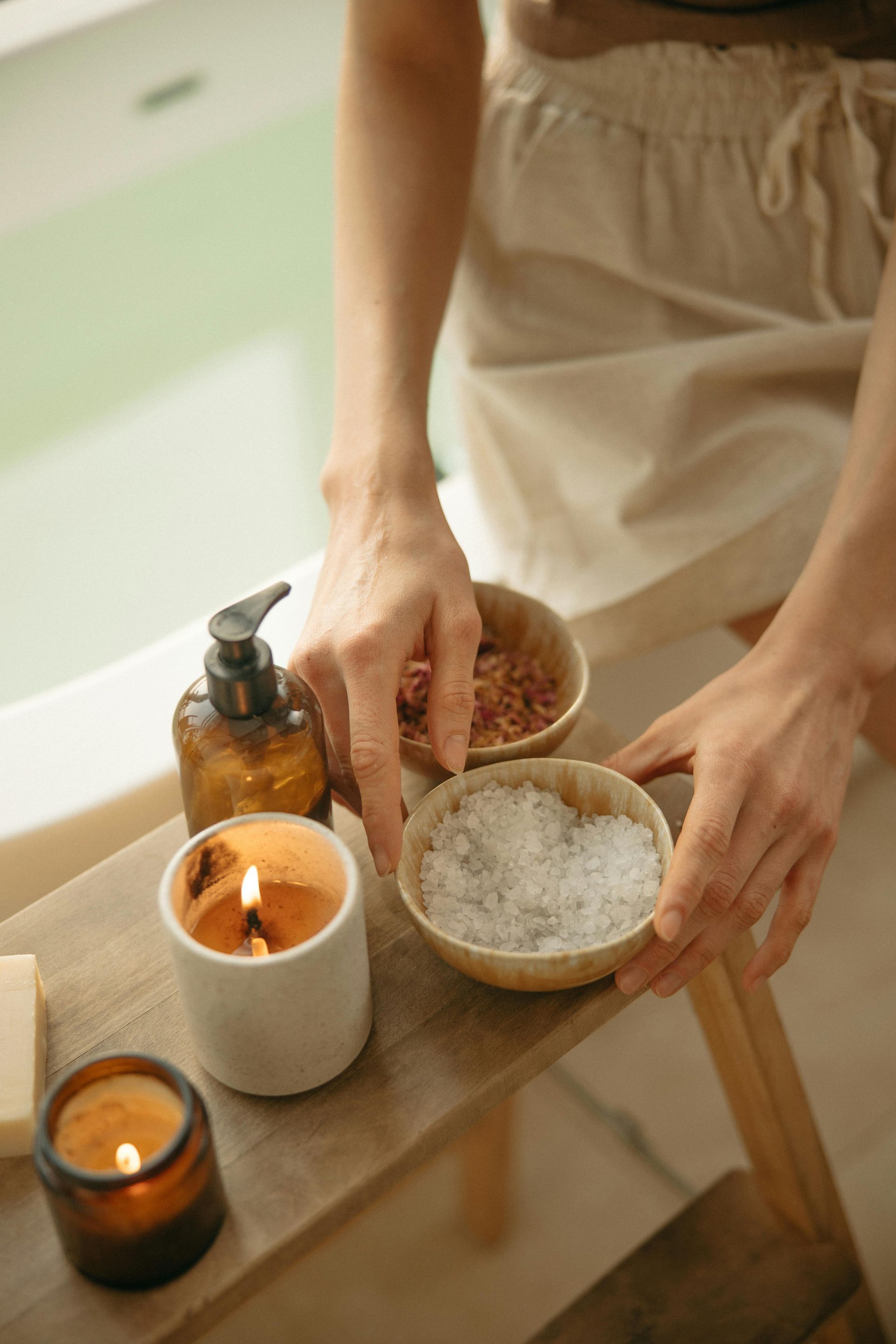 Person's hands with bath salts, candles, and lotion on a wooden shelf near a bathtub.