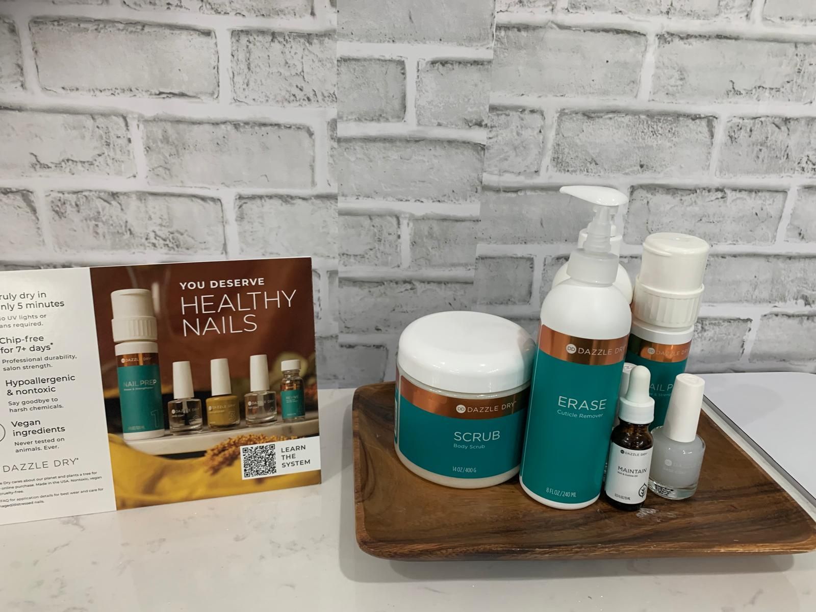Nail care products arranged on a wooden tray, with a brochure and a brick wall backdrop.