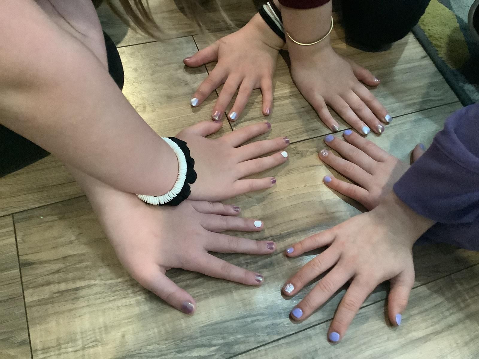 Four hands with painted nails forming a circle on a wooden floor, some wearing bracelets.