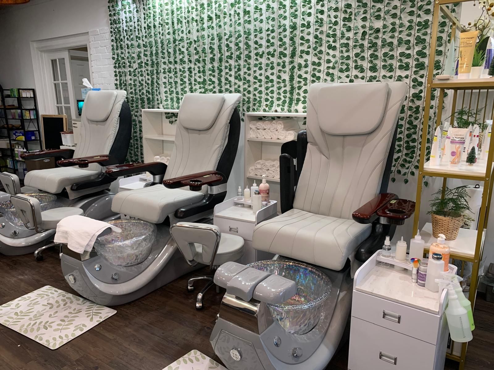 Nail salon with three pedicure chairs. Light gray chairs, foot basins, shelves with products, and a patterned wall.
