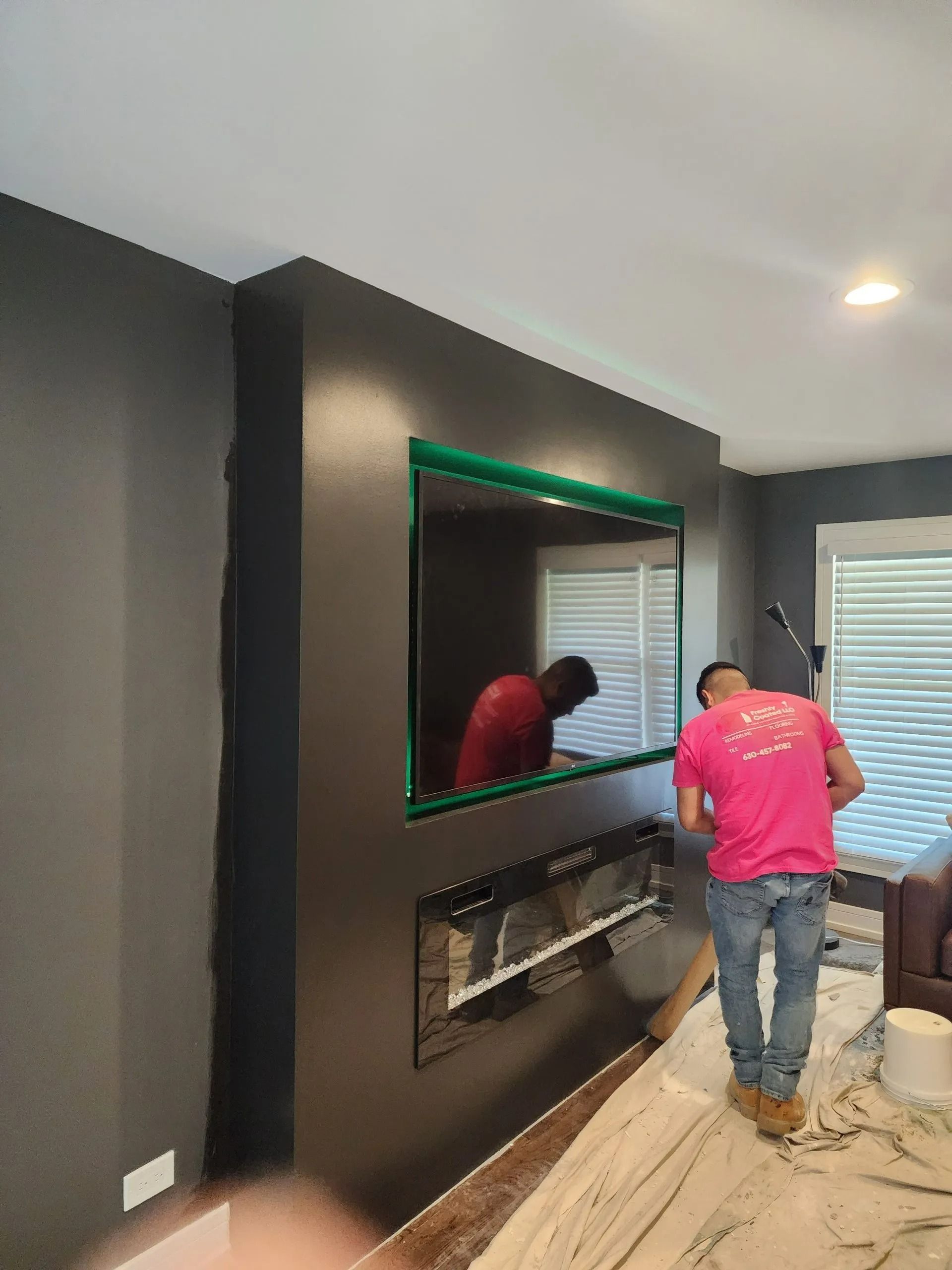 Men install a fireplace under a large TV, on a dark gray wall. Green tape outlines the TV.