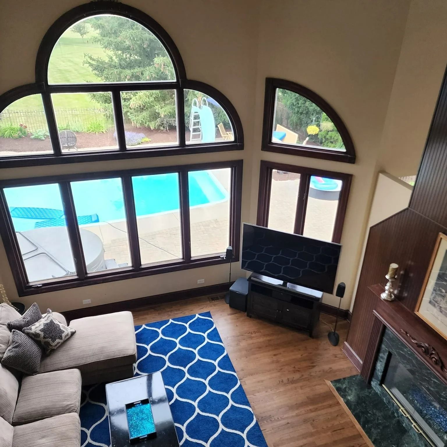 High-angle view of living room with large windows overlooking a pool. Tan couch, blue rug, TV, and fireplace are visible.