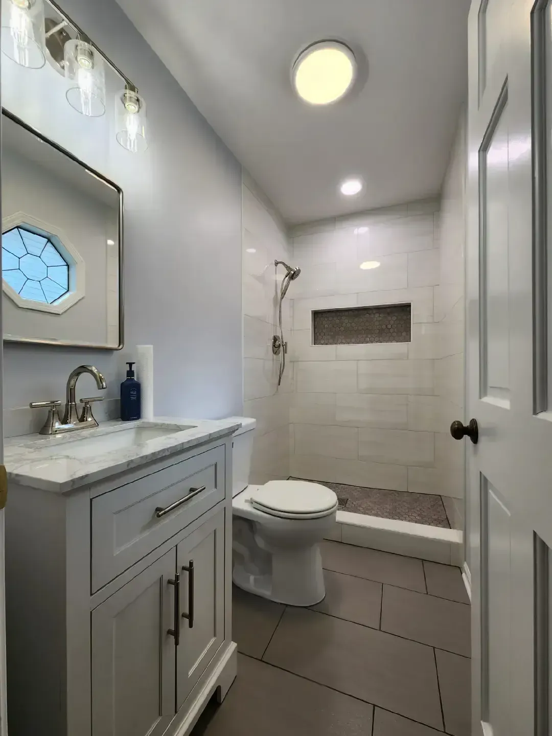 Small, modern bathroom with white vanity, tiled shower, and toilet. Light gray walls and floor.