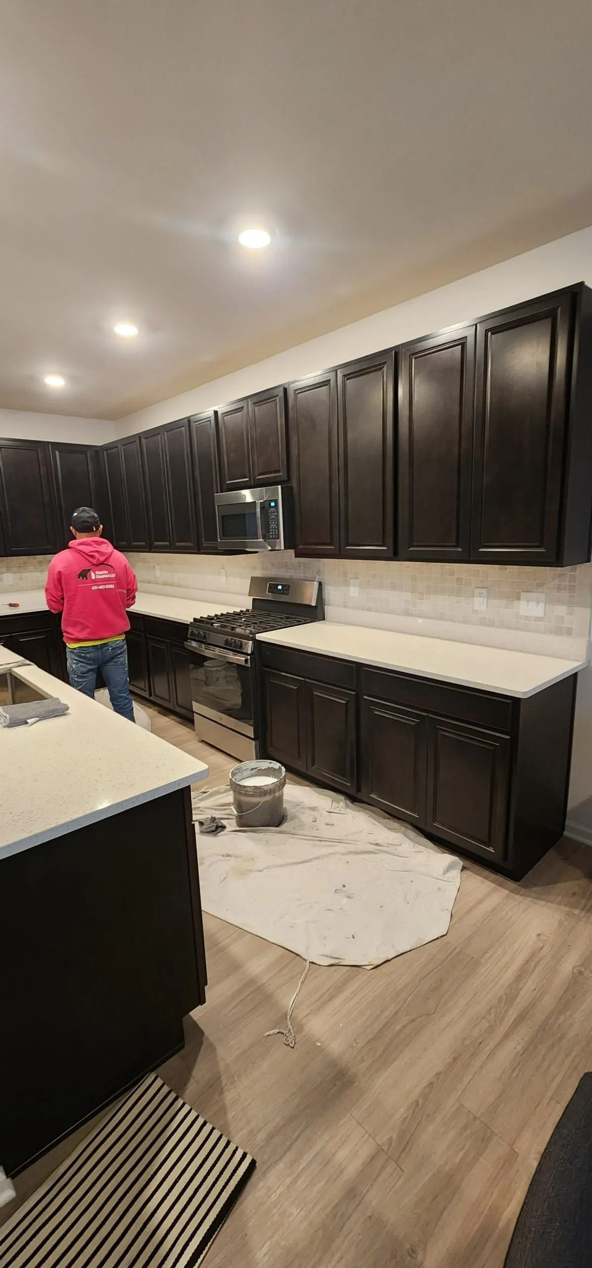A kitchen with dark cabinets, light countertops, and a person in a pink shirt.