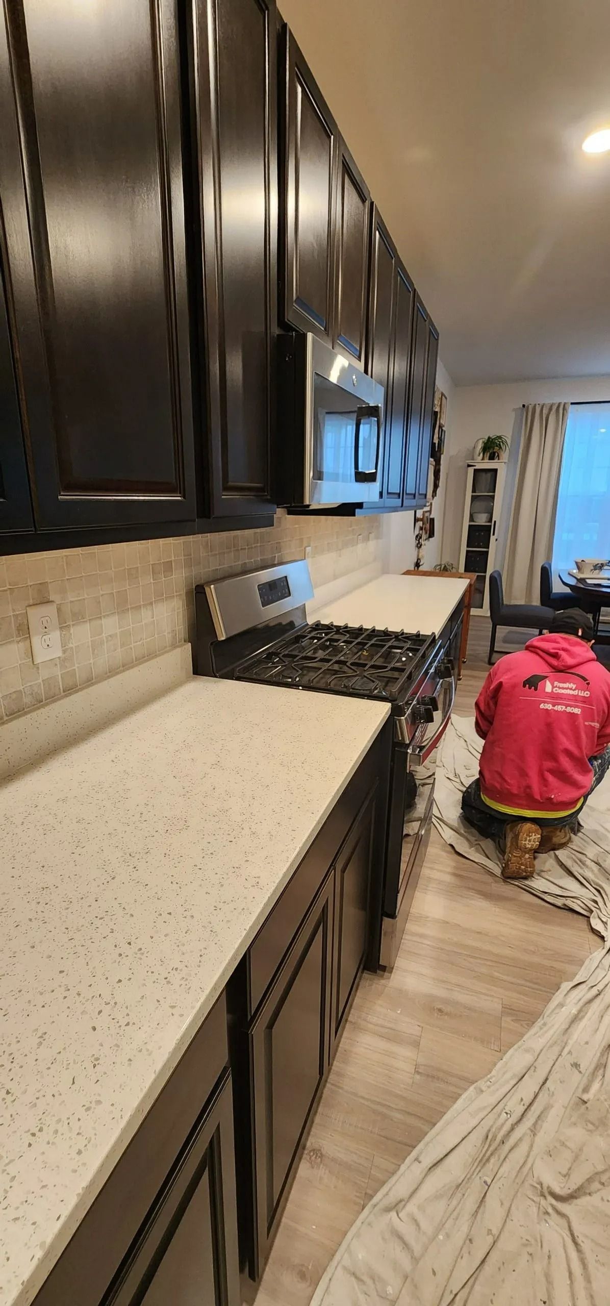 A kitchen with dark cabinets, light countertops, and a person in a red shirt working on the floor.