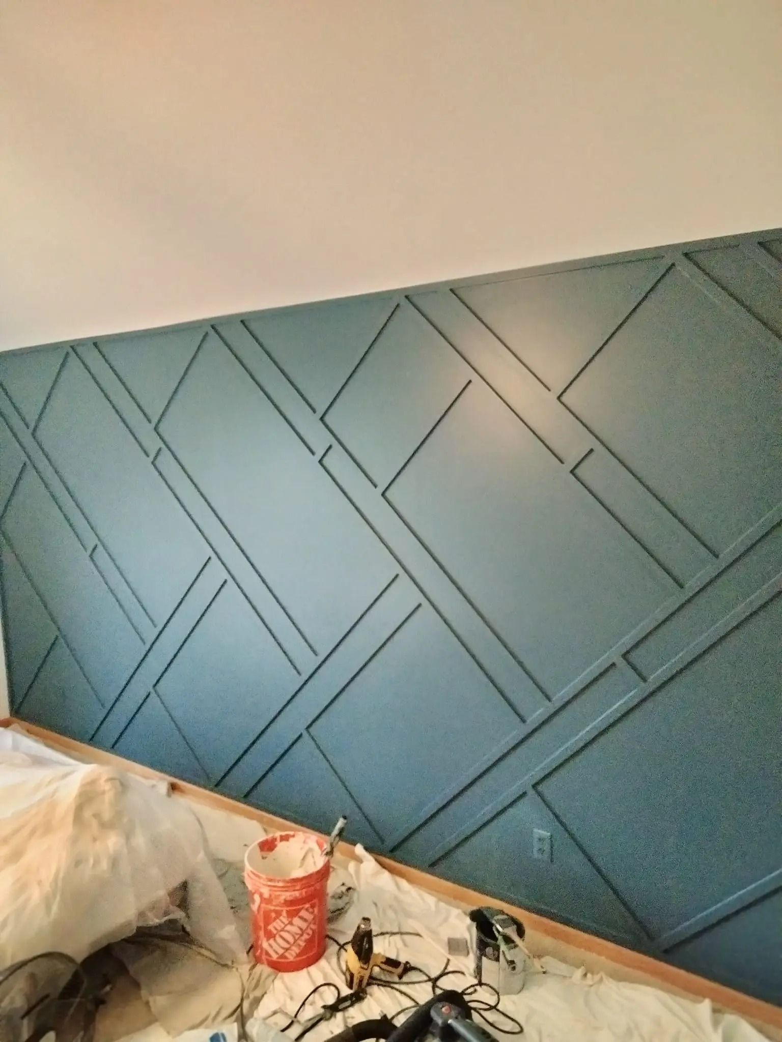 Teal accent wall with geometric wood paneling. A paint bucket and tools sit on the floor.