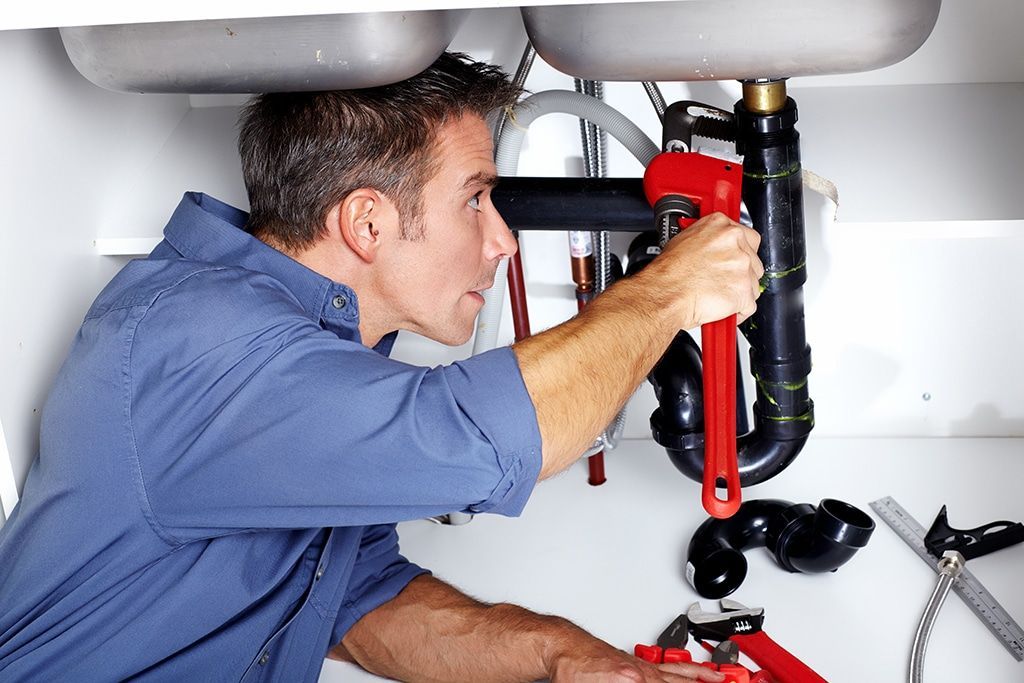 Plumber using a red wrench to repair pipes under a kitchen sink.