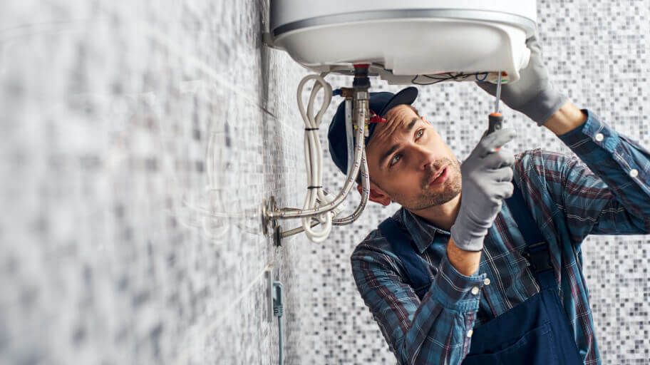 Plumber fixing a water heater in a tiled bathroom, using a screwdriver.