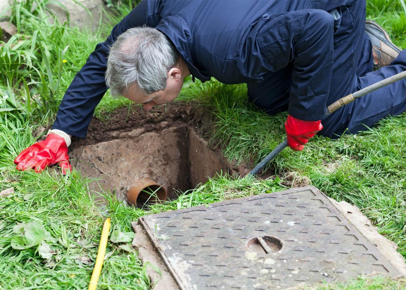 Man in blue jacket, red gloves, inspecting a pipe in a hole near a manhole cover in grass.