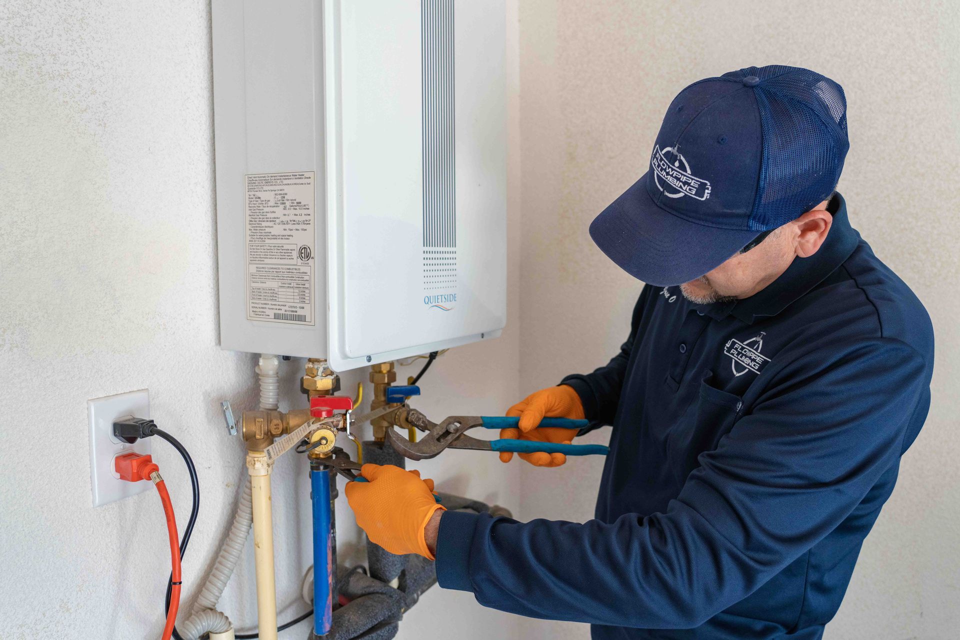 Plumber in a blue uniform and cap using pliers to work on a wall-mounted water heater near electrical outlets.