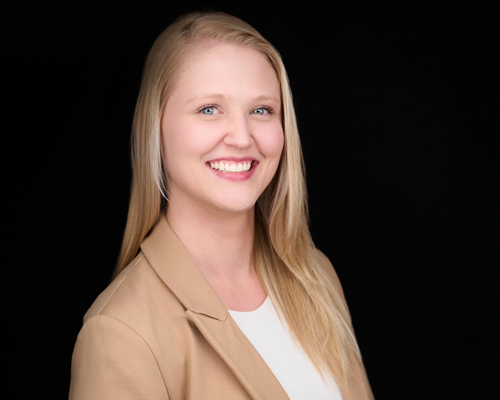Woman with long blonde hair, smiling, wearing a tan blazer against a black background.