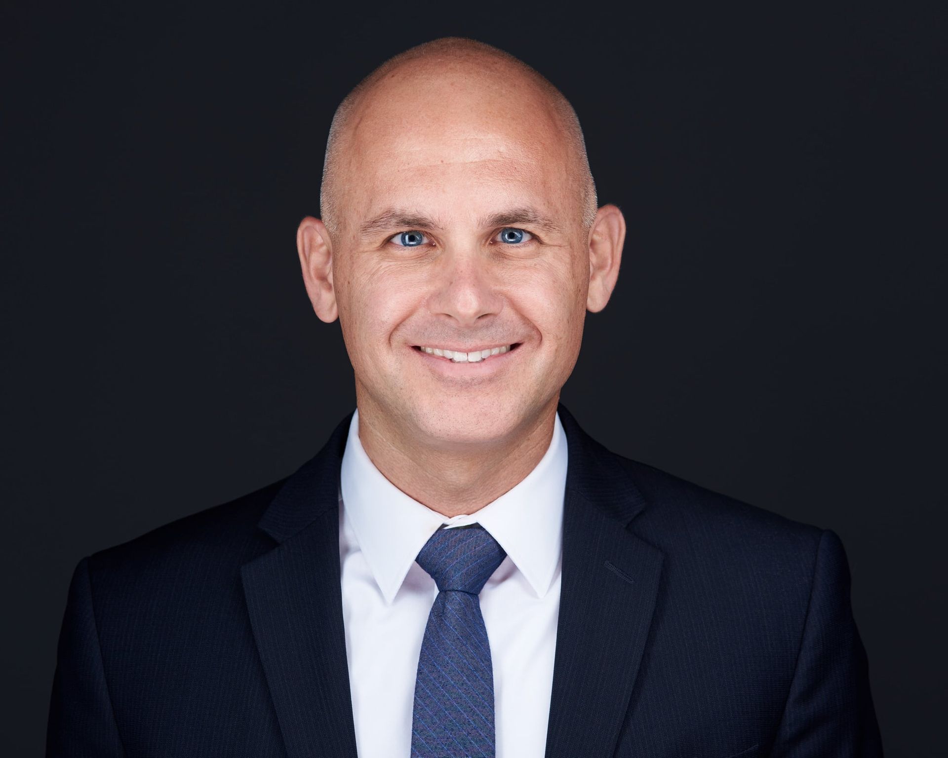 Bald man in a suit smiling at the camera against a dark background.