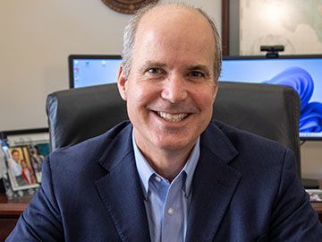A man in a suit is smiling while sitting in a chair in front of two computer monitors.