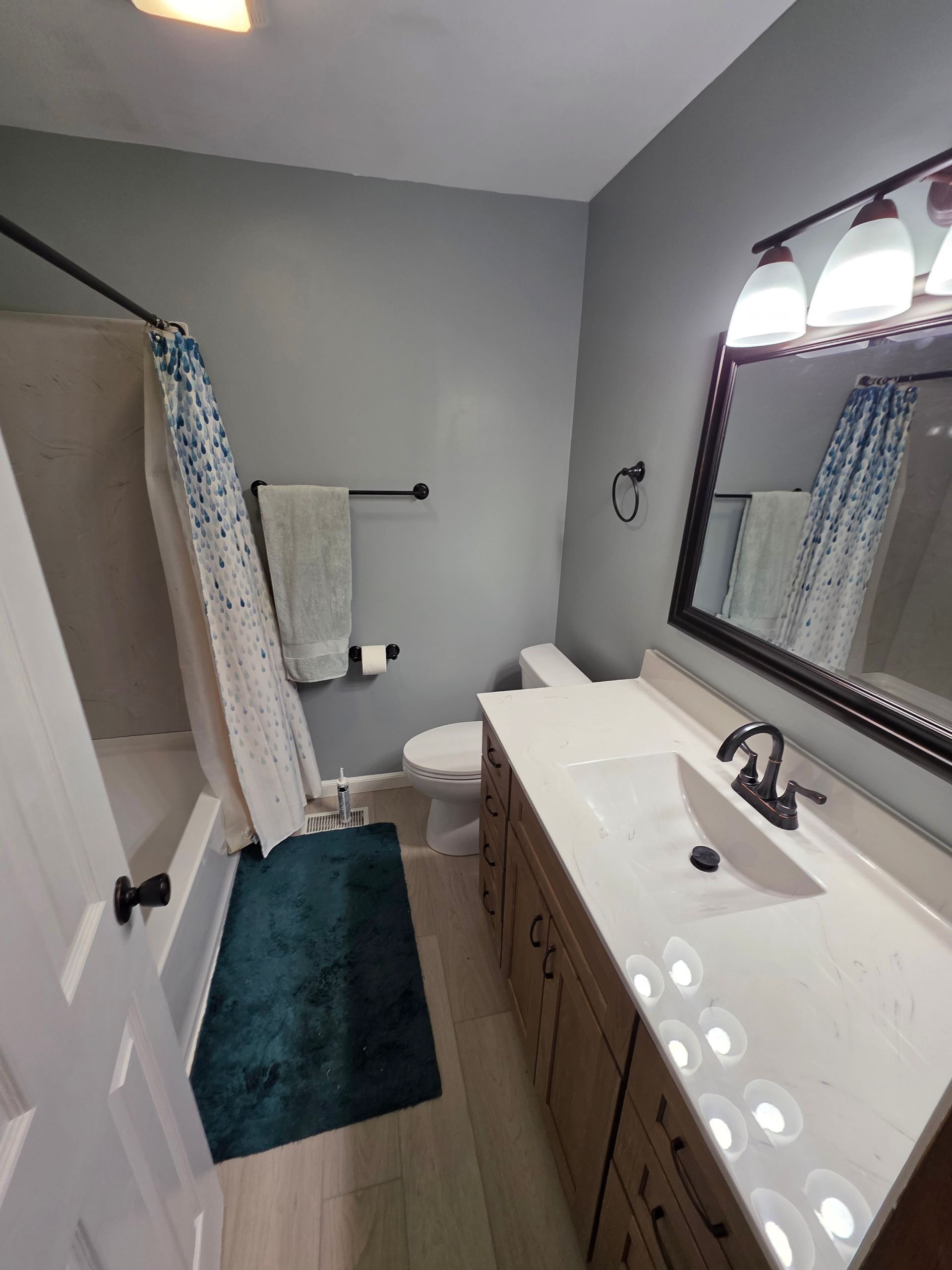 Bathroom with gray walls, white countertop, wooden cabinets, and teal shower curtain and rug. | Danville, IL