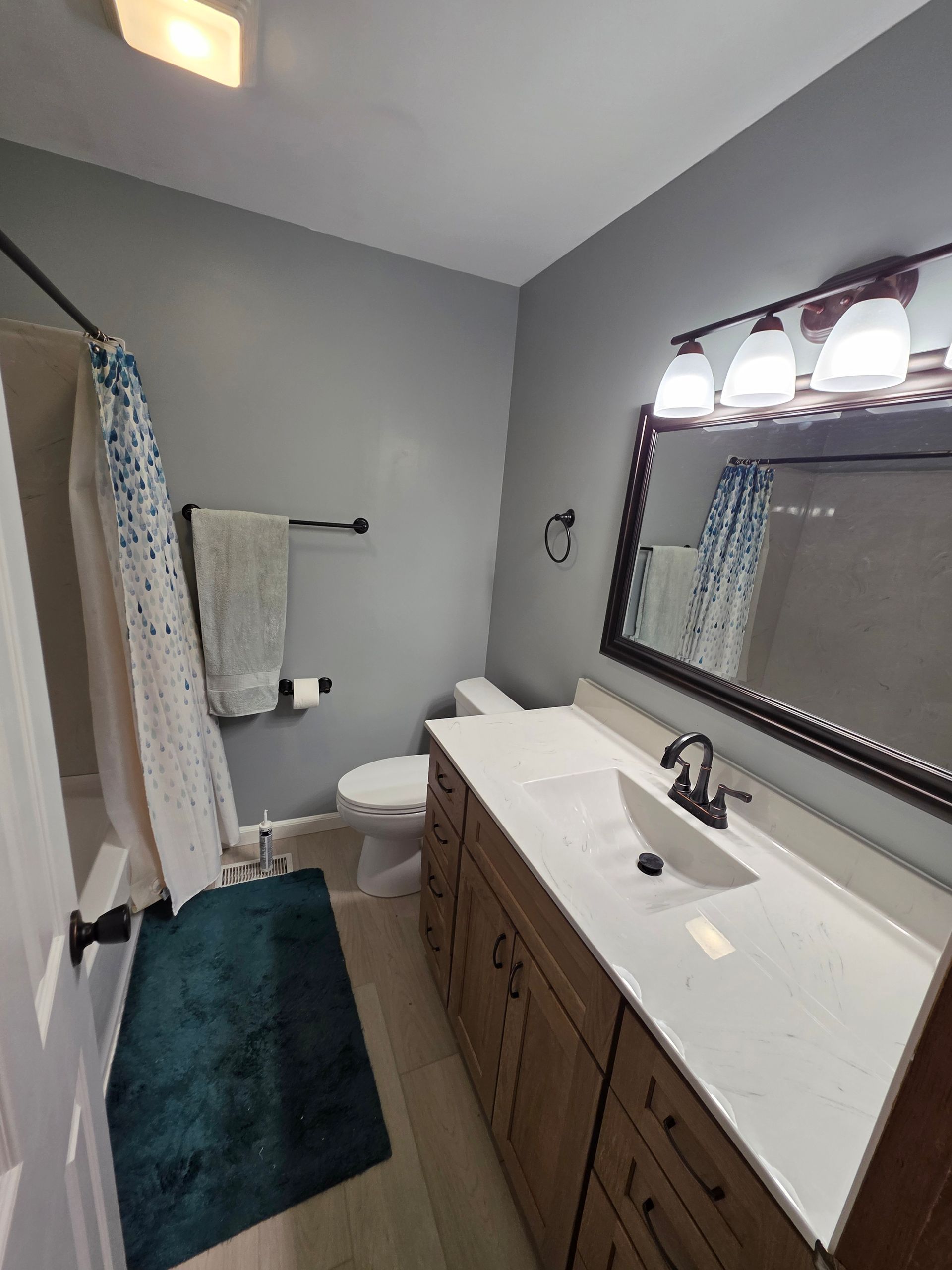 Bathroom with a blue shower curtain, teal rug, gray walls, and a wooden vanity.| Danville, IL