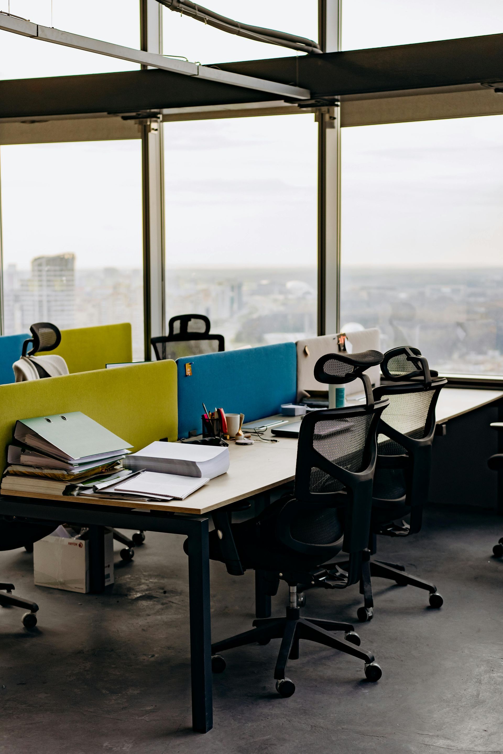 Office cubicles with desks and chairs, facing a large window overlooking a cityscape.