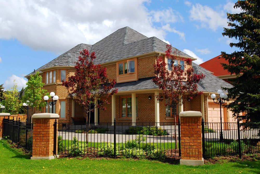 Two-story tan brick house with grey roof and black iron fence.