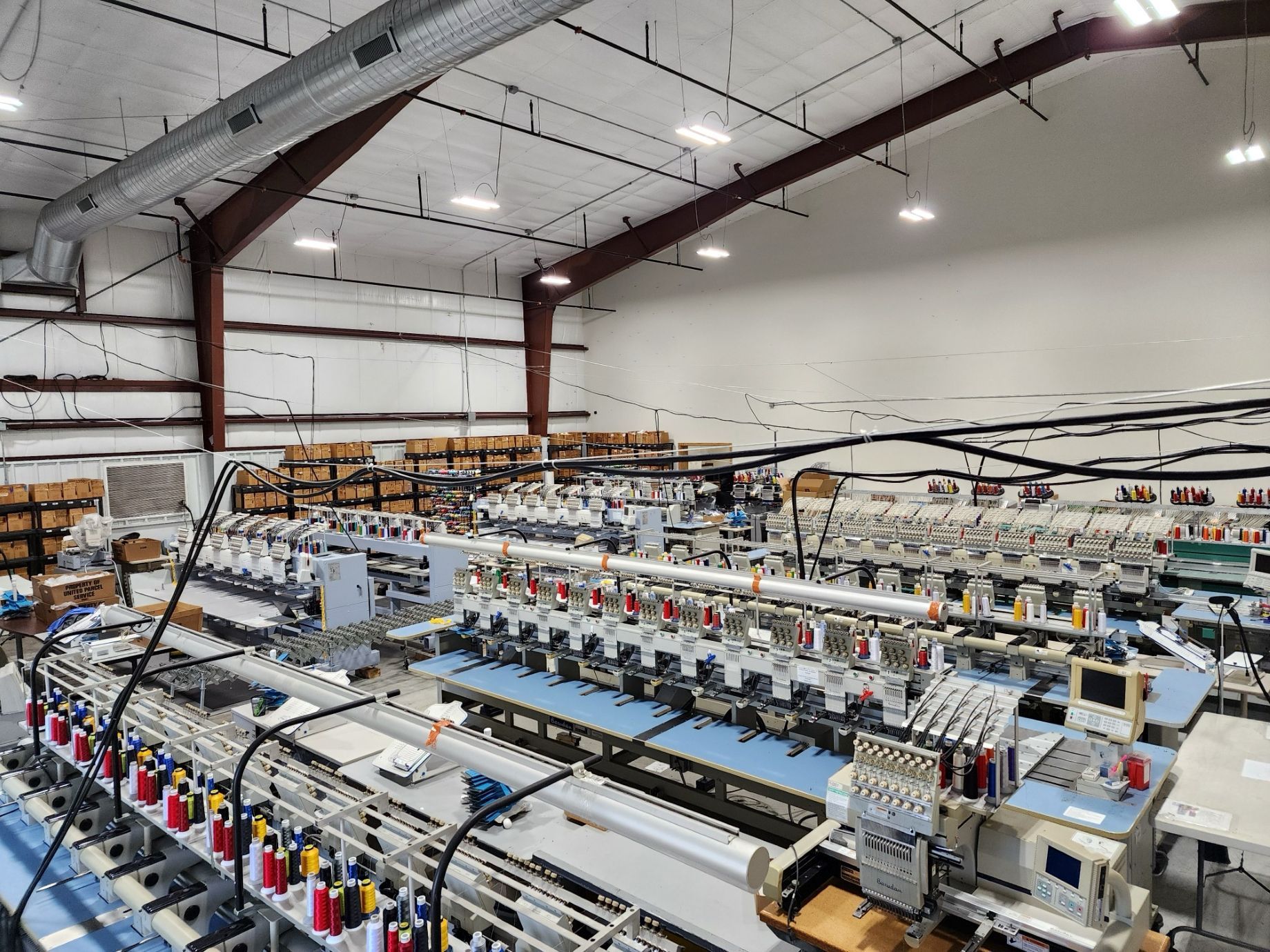 Embroidery machines in a large factory setting. Brown and white building with boxes on shelves in the background.