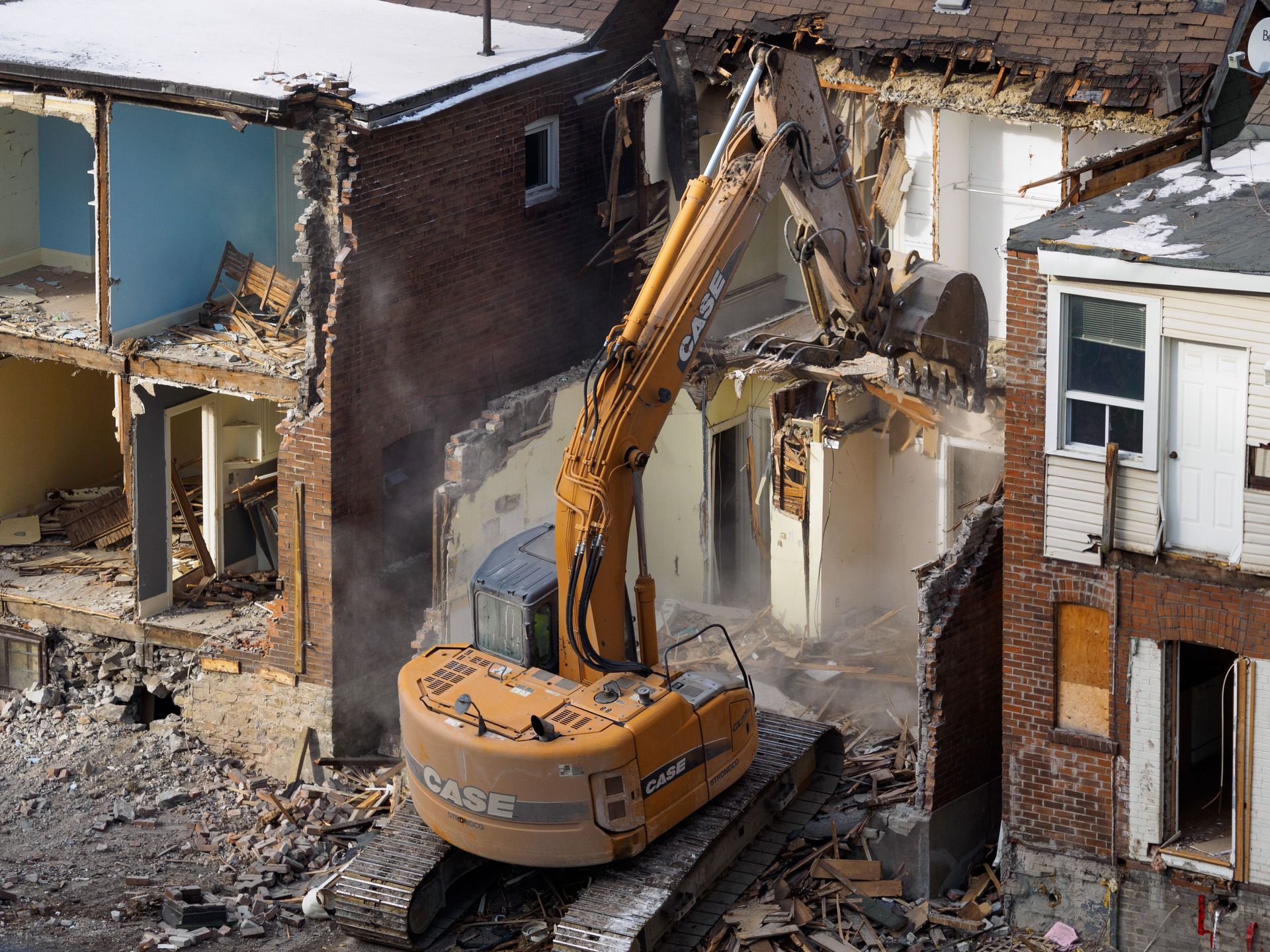 Excavator demolishing a damaged house in a row of homes, with rubble and exposed walls.