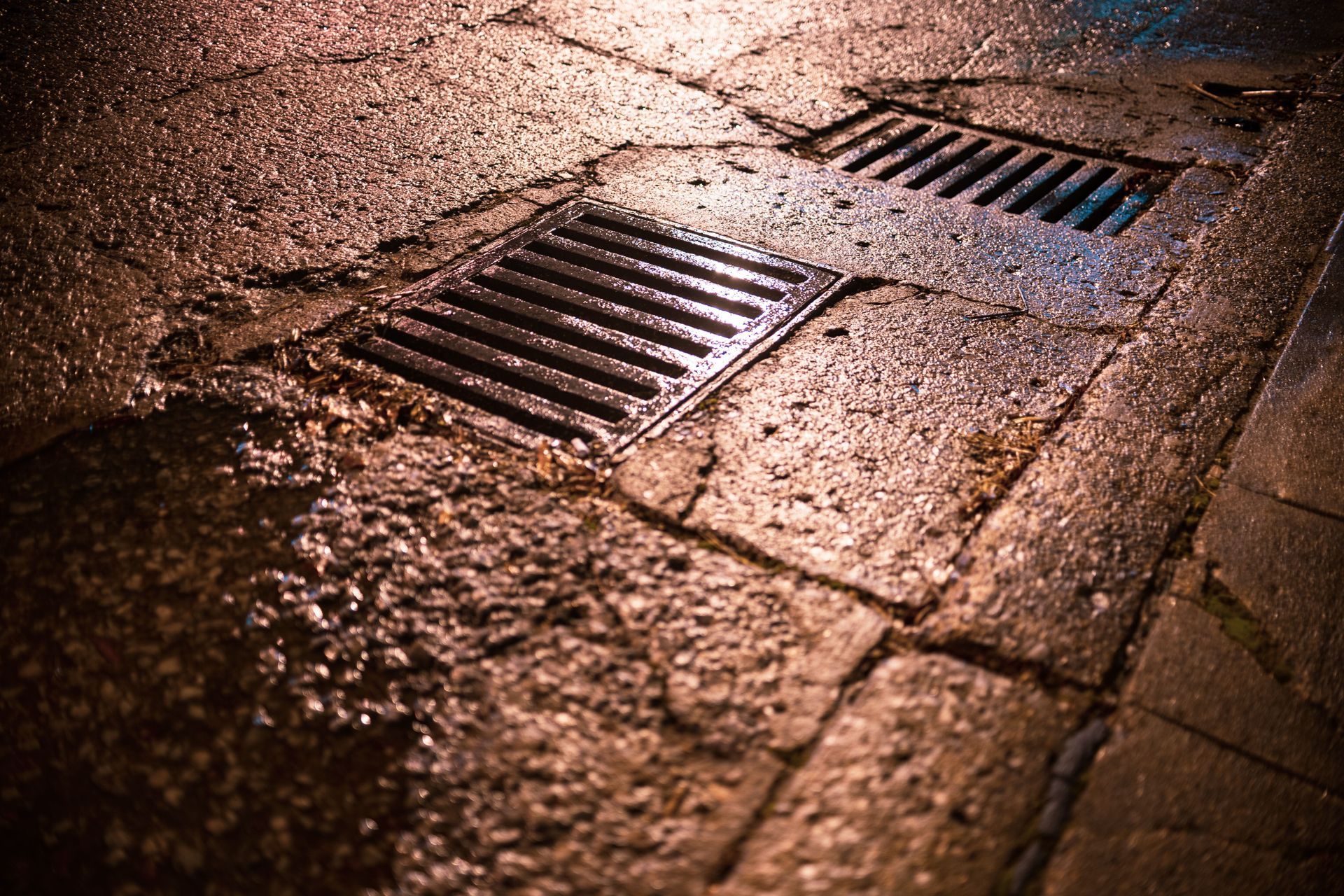 Wet cobblestone pavement with two metal storm drains at night, lit by warm streetlight reflections