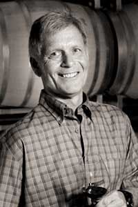 Man smiling in plaid shirt, barrels in background.