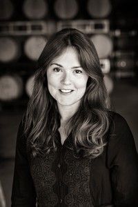 Woman with long hair smiles, wearing a dark blouse, in front of wine barrels.