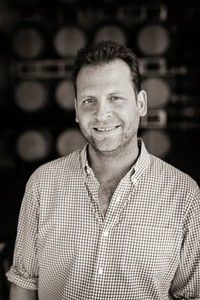Man in checkered shirt smiling, standing in front of blurred wooden barrels.