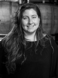 Woman with long hair smiles in front of wooden barrels, wearing a black top.