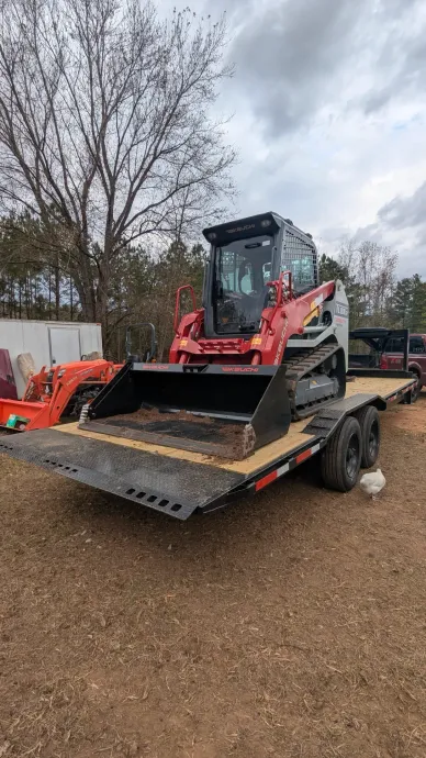 Red skid steer loader on a flatbed trailer in a gravel lot, with trees and overcast sky behind it