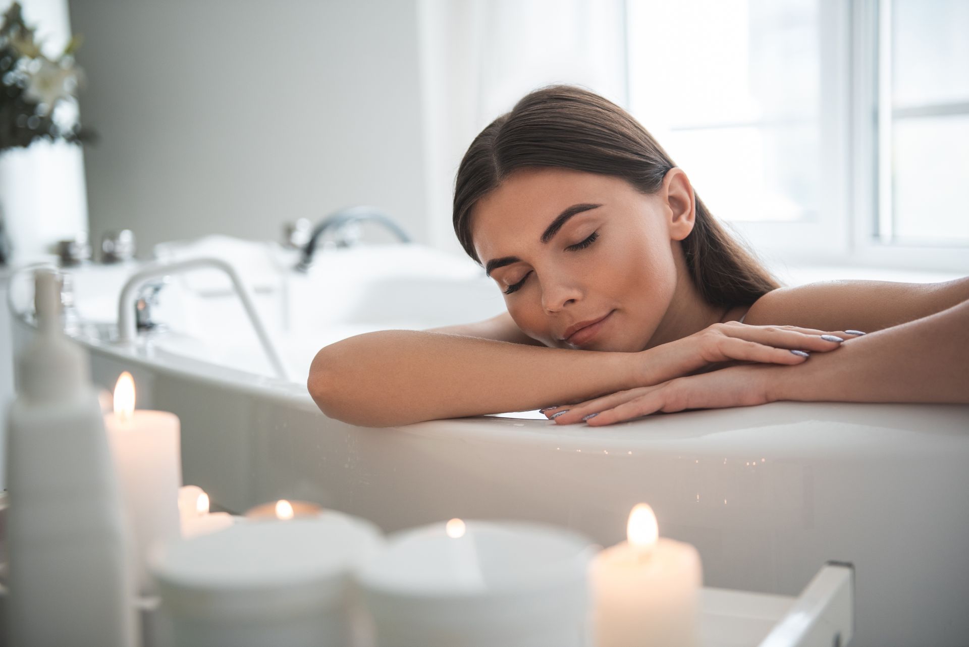 A serene woman leaning on the side of a bath while resting with candles in the foreground.