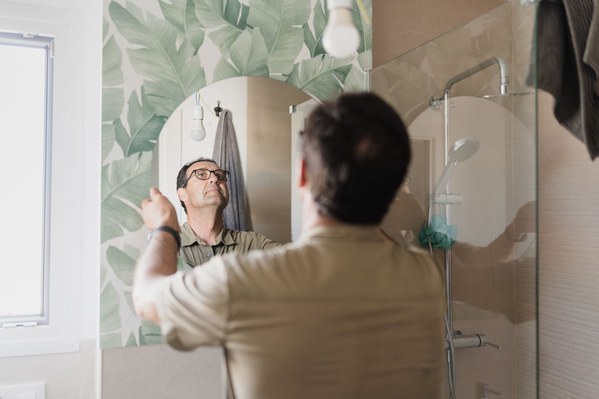 Person pointing at round mirror in bathroom with leafy wallpaper.