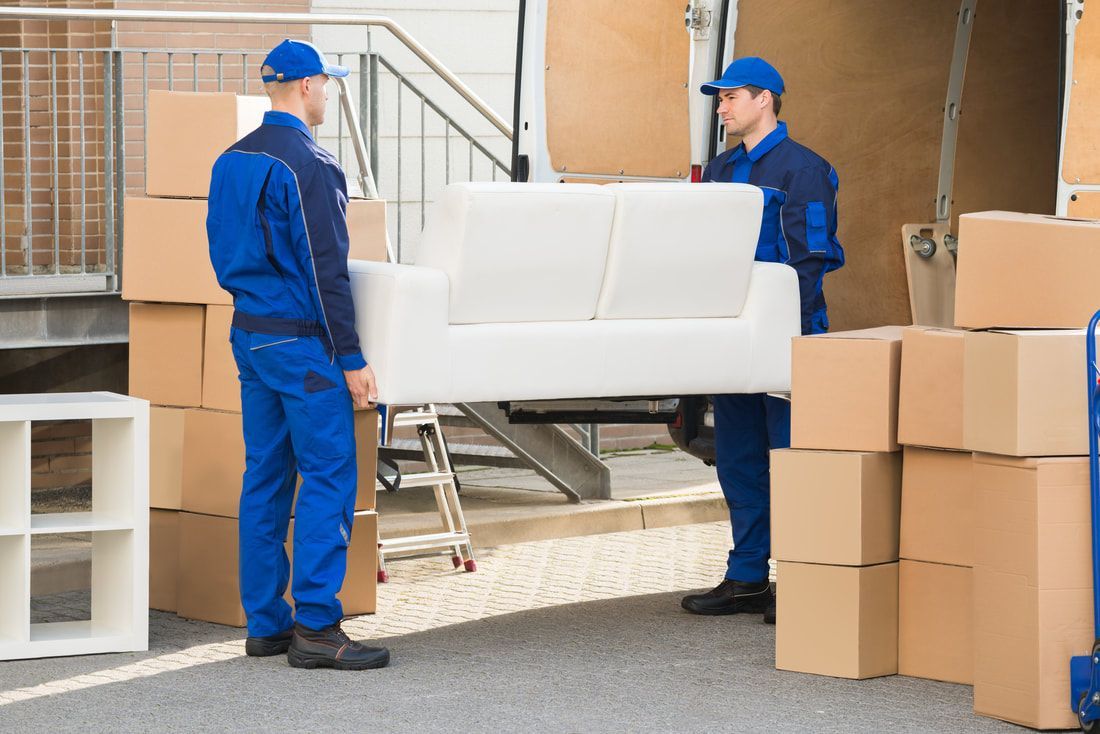 Two movers in blue uniforms lift a white couch from a truck, surrounded by cardboard boxes.