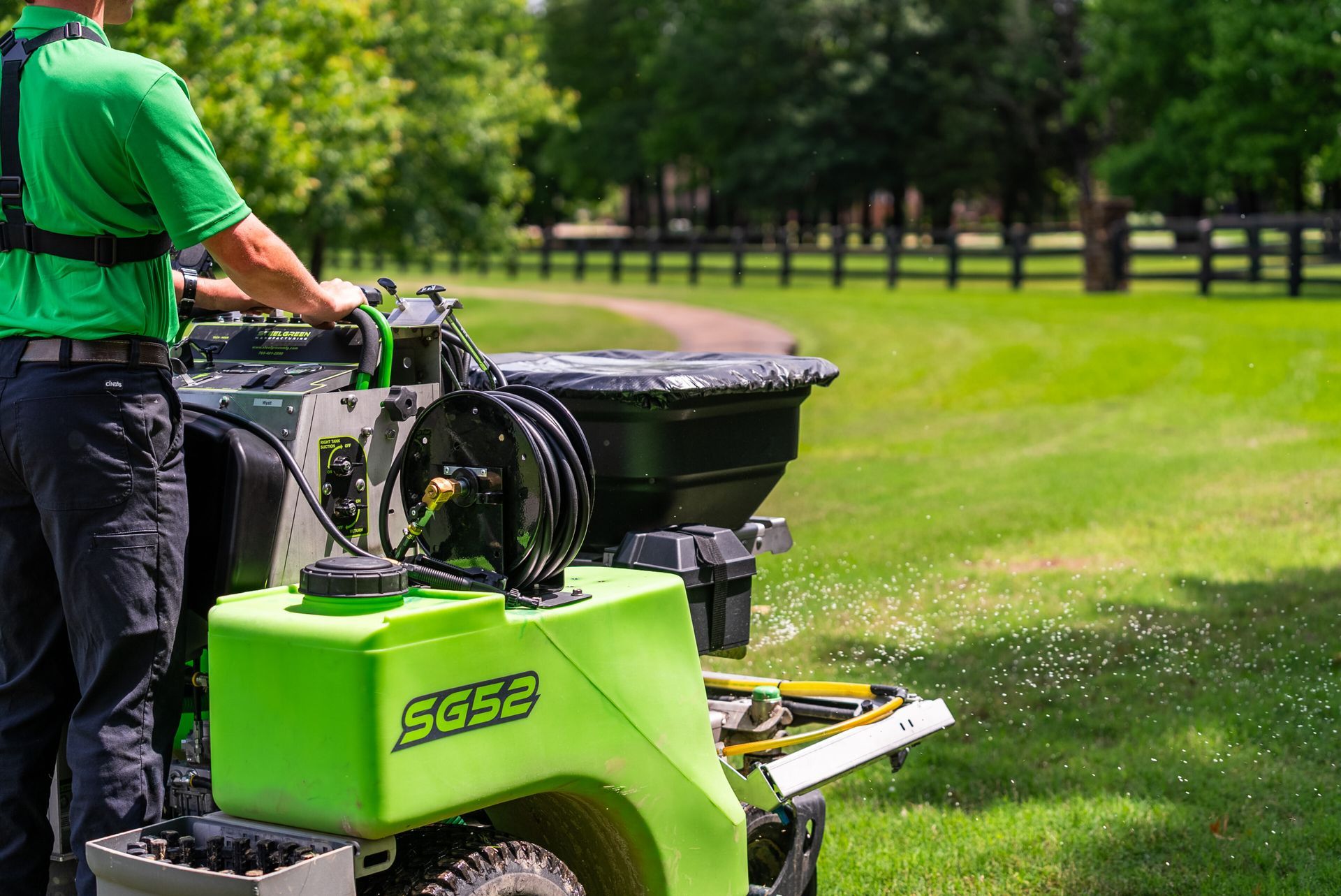 Man in green shirt operating a green SG52 machine spraying a lawn. Outdoors with a fence in the background.