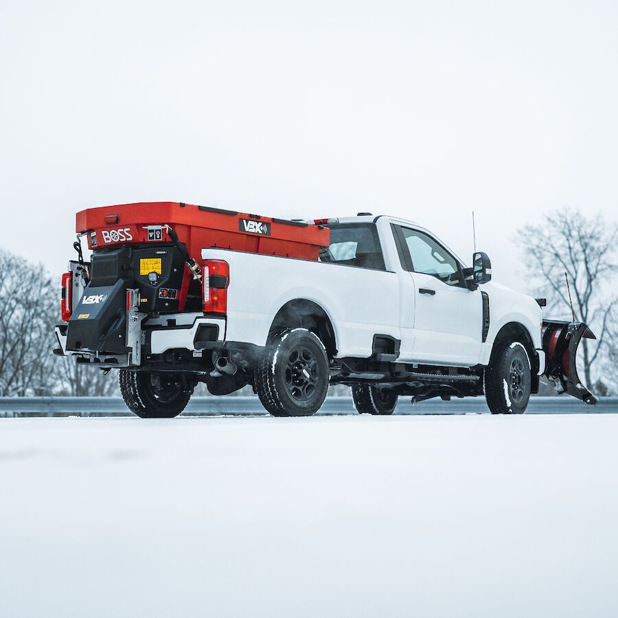 White pickup truck with snowplow and salt spreader on a snow-covered road.