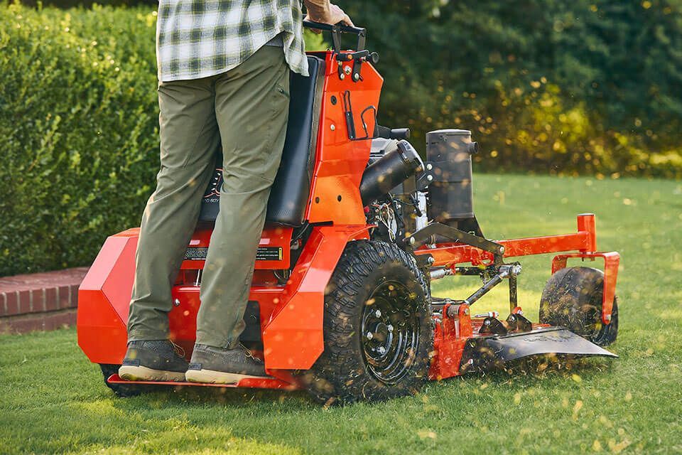 Person operating an orange stand-on mower on a grassy lawn. Green plants and brick wall in the background.