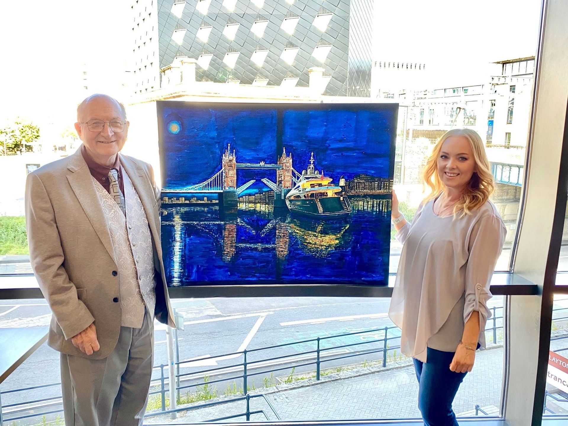 image of renowned Welsh Artist Jeffrey Phillips with his painting of a Superyacht at Tower Bridge, London. Pictured is agent Donna Phillips